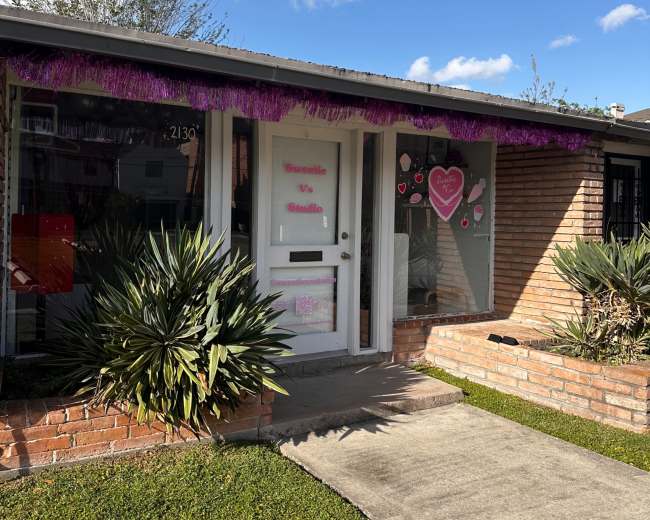 The image shows a small brick building with a front door adorned with decorations, including hearts and a purple fringe, set against a clear blue sky.