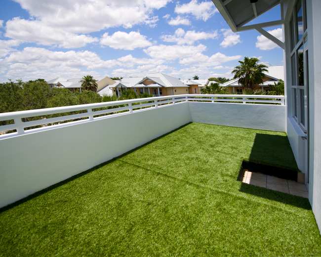 The image shows a spacious balcony featuring artificial grass, surrounded by a white railing and overlooking residential buildings beneath a partly cloudy sky.