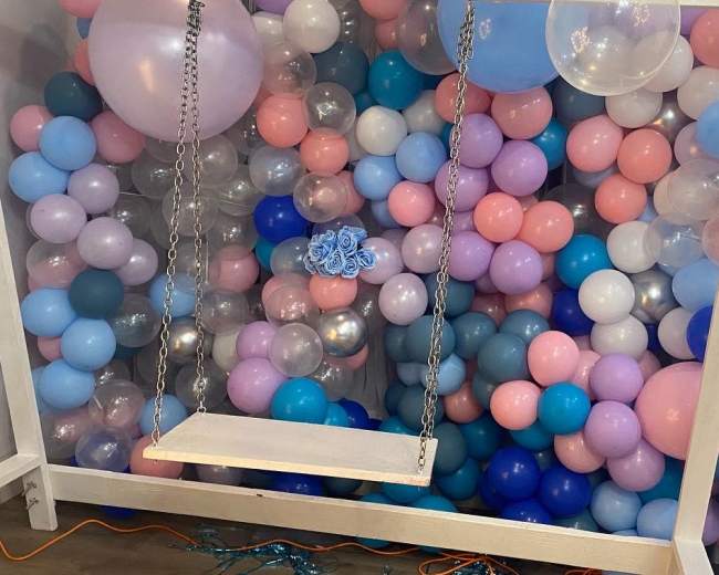 A wooden swing is positioned in front of a colorful backdrop made of assorted balloons in shades of pink, blue, and silver.