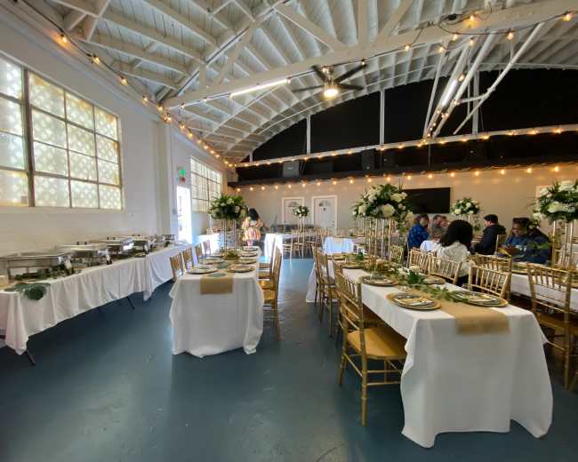 A large event space is arranged with gold chairs and tables set for a formal gathering, featuring buffet stations and floral decorations.