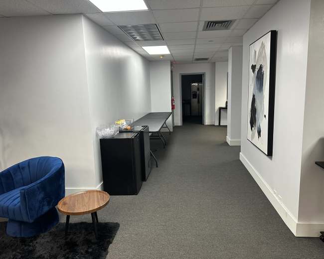 The image shows a hallway with gray carpeting, a blue chair and a small wooden table to the left, and a door at the end leading to another room.