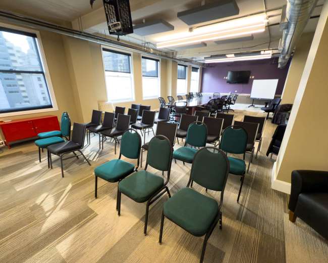 The image shows a conference room with rows of green chairs set up facing a presentation area, accompanied by a few tables and a whiteboard.