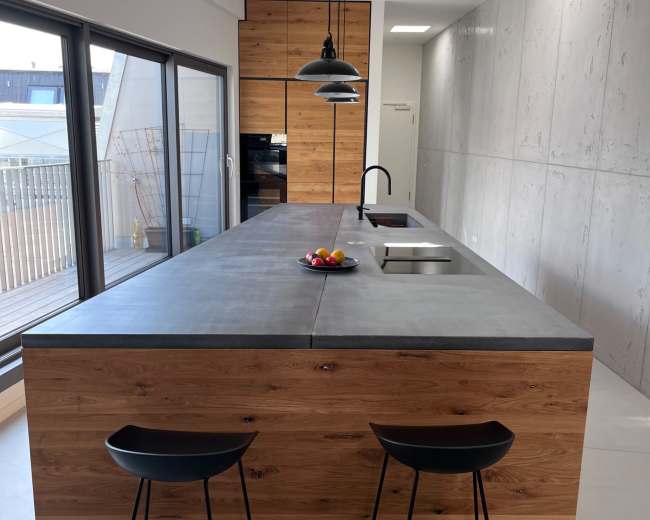 The image shows a modern kitchen with a large concrete countertop, wooden cabinetry, and two black stools.