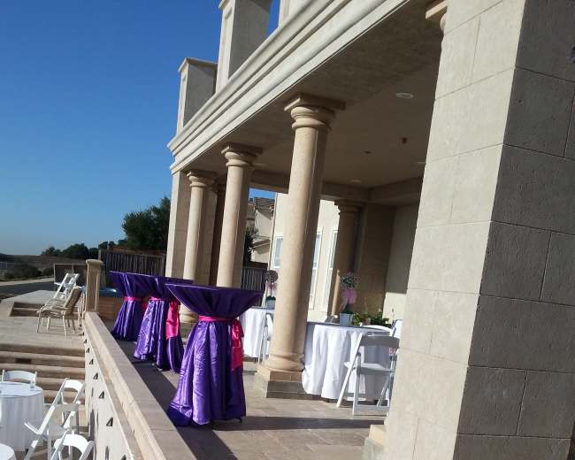 The image shows a patio area with columns, featuring tables draped in purple and pink tablecloths, set up for an event.