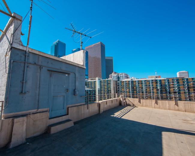 The image shows a rooftop area with a concrete structure and several buildings in the background, under a clear blue sky.