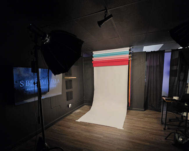 The image shows a photography studio with a white backdrop, colored paper rolls, softbox lights, and a viewing screen in a dimly lit room.
