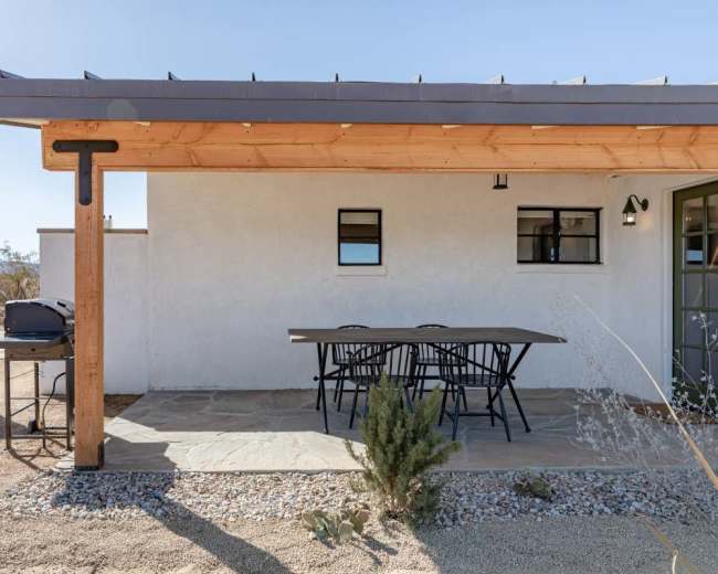 The image shows a modern outdoor patio area with a wooden overhang, a dining table with black chairs, and a grill next to a desert landscape.