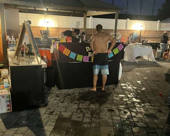 A shirtless man stands in front of a decorated bar counter under a gazebo at a nighttime outdoor event.