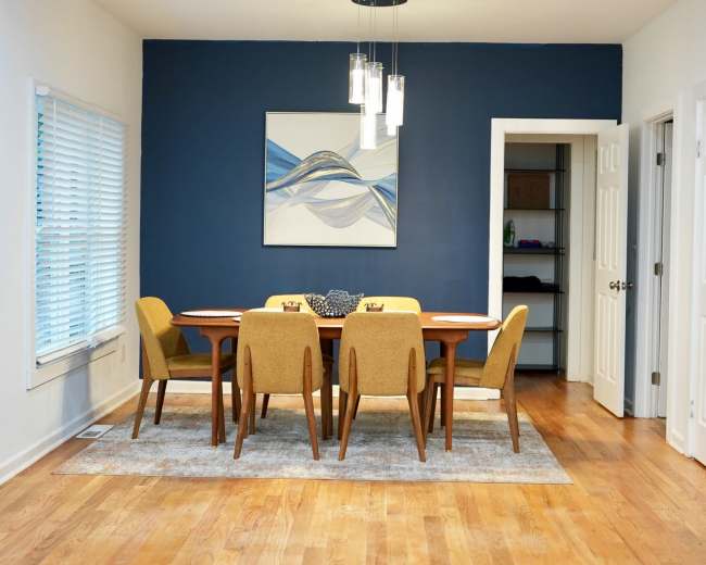 A modern dining room features a rectangular table surrounded by yellow chairs, with a large abstract painting on a blue accent wall.