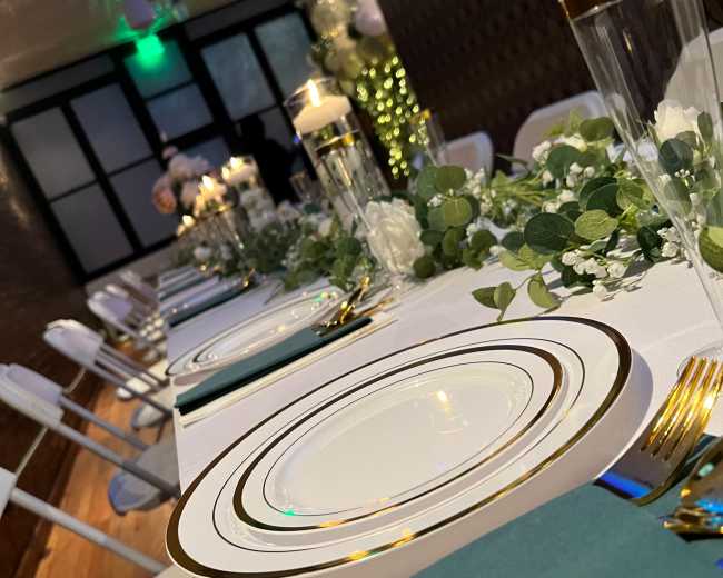 A long table is elegantly set with white plates featuring gold trim, teal napkins, and greenery decorations.