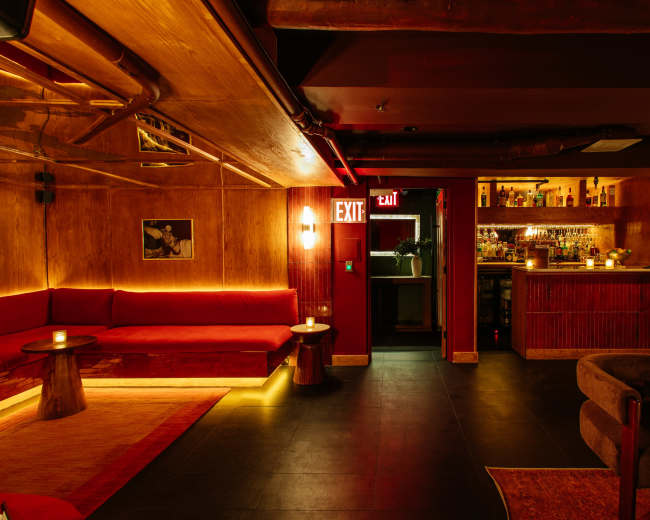 The image shows a dimly lit bar interior featuring red seating, wooden paneling, and a well-stocked bar area in the background.