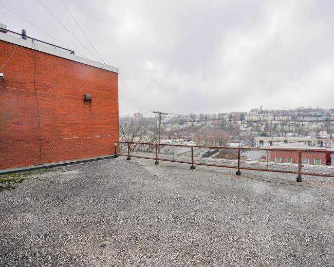 The image shows a rooftop area with a gravel surface and a railing overlooking a hillside populated with buildings and trees under a cloudy sky.