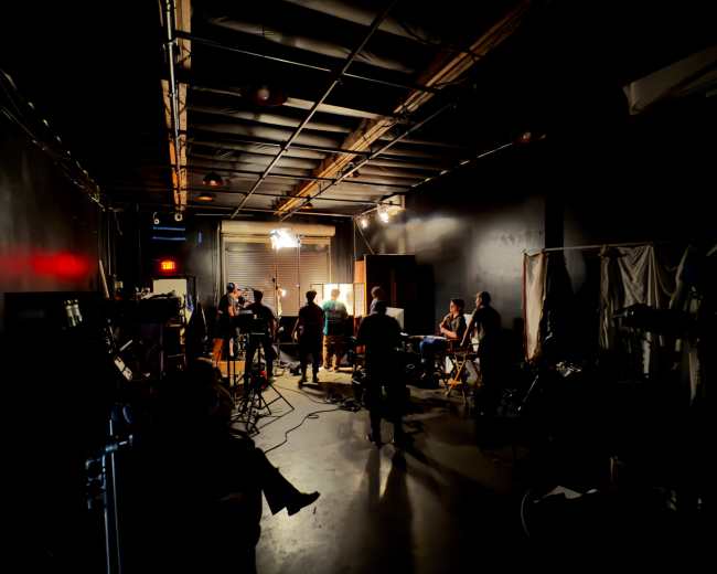 A film crew works on a set in a dimly lit studio, surrounded by equipment and various tools.