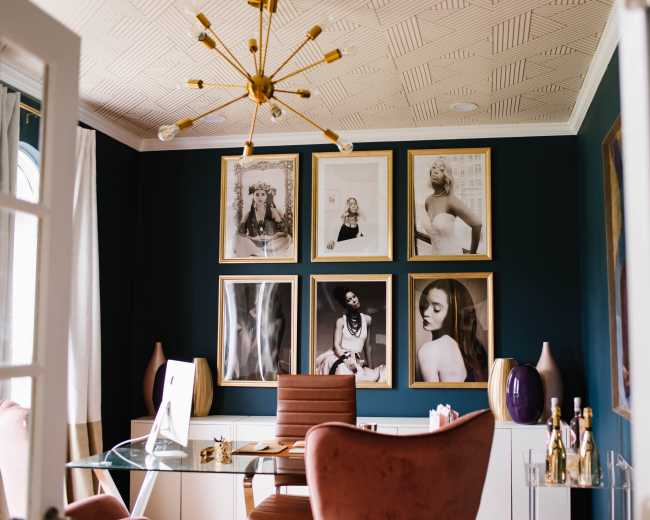 The image shows an elegant home office with a dark green wall adorned with framed black-and-white photographs, a modern desk, and a stylish chair.