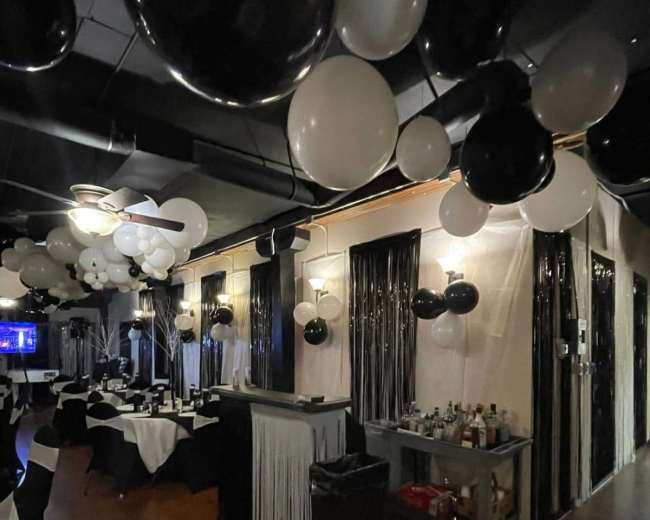 The image shows a decorated restaurant interior with black and white balloons hanging from the ceiling and tables set for dining.