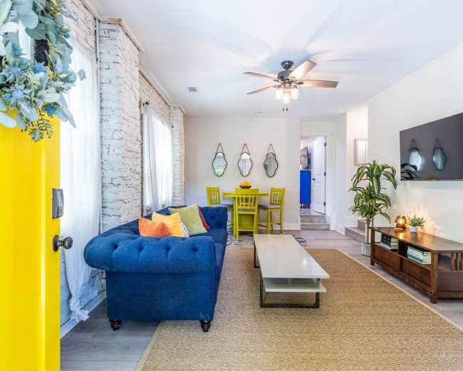 The image shows a modern living room with a blue tufted couch, a coffee table, a television mounted on the wall, and bright yellow accents.