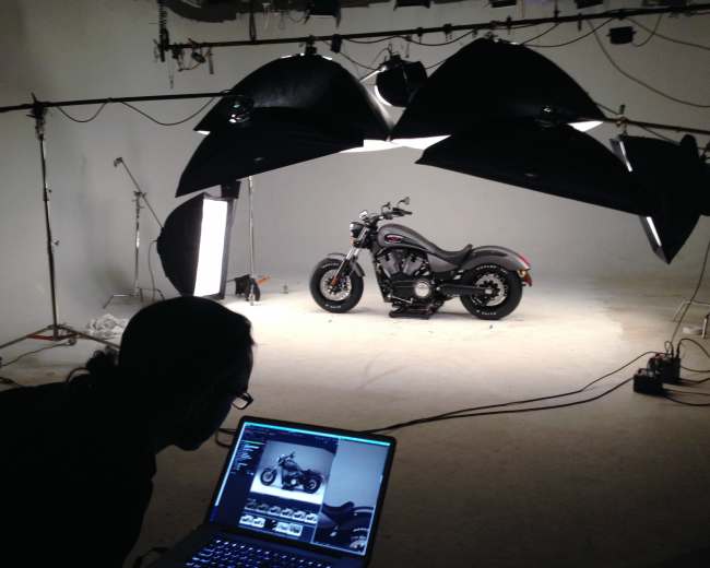 A motorcycle is displayed under studio lighting while a person works on a laptop in the foreground.