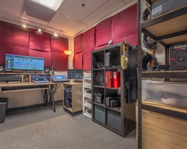 A small studio room features equipment for music production, including a computer setup, shelves with gear, and soundproofing panels on the walls.
