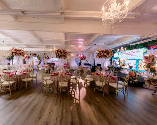 The image shows a beautifully decorated event space with round tables set for a celebration, adorned with floral centerpieces and lavish decorations.