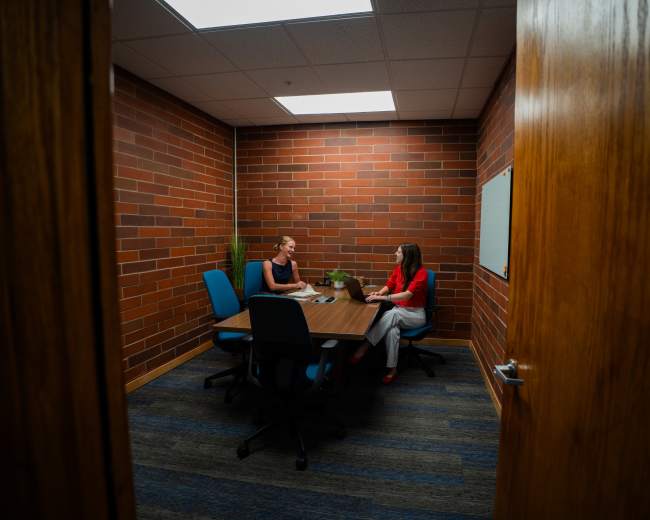 Two women are seated at a conference table in a small, brick-walled meeting room.