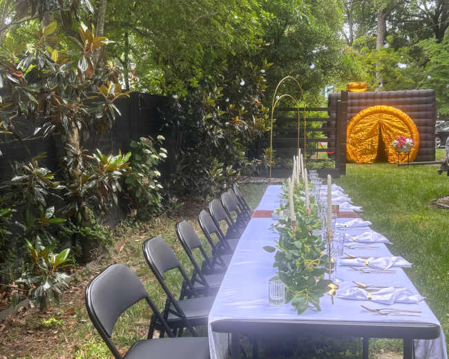 A long, neatly arranged table set for a gathering is placed outdoors among greenery, with black folding chairs lining one side and a colorful decoration in the background.