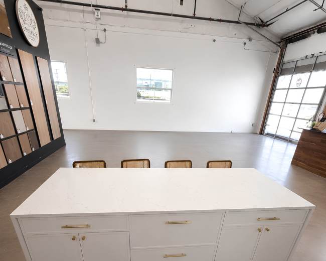 The image shows a white countertop with gold handles, positioned in a well-lit, open space with four chairs in the background.