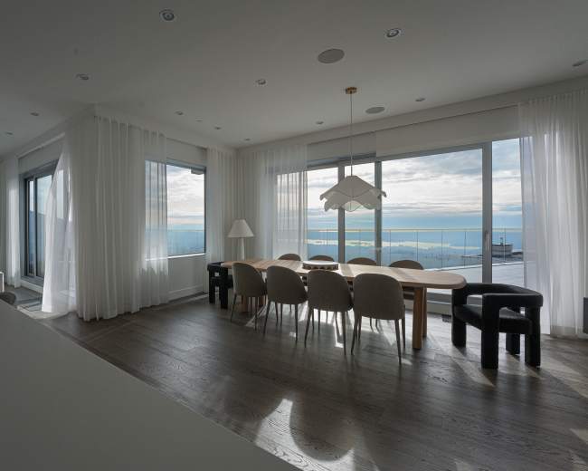 A spacious dining area features a large wooden table surrounded by upholstered chairs, with large windows offering a view of the water and sky.