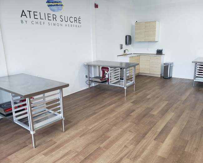 The image shows a bright, minimally furnished kitchen space with stainless steel tables and a wooden floor, featuring a kitchenette in the background.