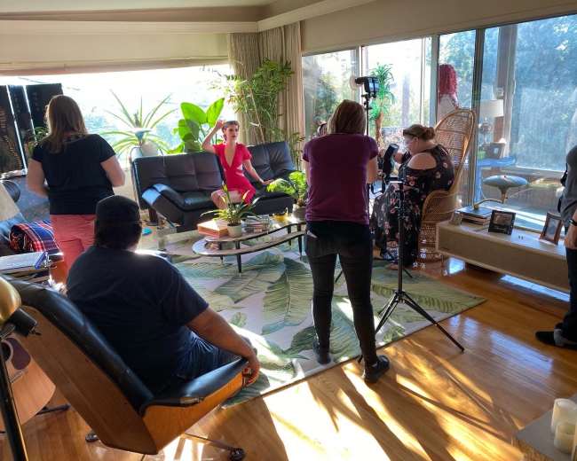 A group of people is gathered in a brightly lit living room, with one person in a red dress posing for a photo while others work with cameras and equipment.