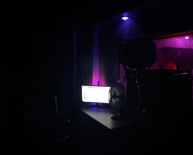 The image shows a dimly lit recording studio with a desk, a computer monitor, a fan, and a microphone setup against a backdrop of soundproofing panels.