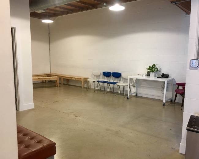 The image shows a spacious, minimalist indoor area with a wooden bench, several chairs, a table, and a potted plant against a white wall.