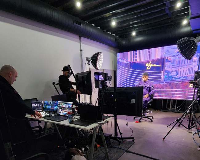A small studio setup features a desk with multiple monitors, two people working behind it, studio lights, and a backdrop displaying a cityscape.