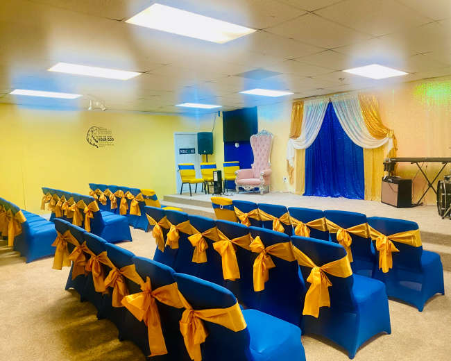 The image shows a brightly decorated event space with rows of blue chairs tied with yellow bows, a stage featuring a pink chair against a backdrop of blue and gold drapes, and a small performance area with musical instruments.
