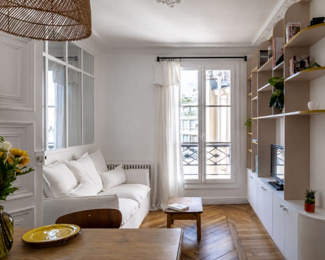 The image shows a bright, compact living room featuring a white sofa, a wooden dining table, and a wall-mounted bookshelf filled with plants and decorative items.
