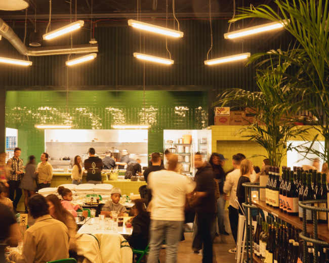 The scene shows a lively restaurant interior with patrons dining at tables, a visible kitchen area with chefs at work, and modern lighting fixtures above.