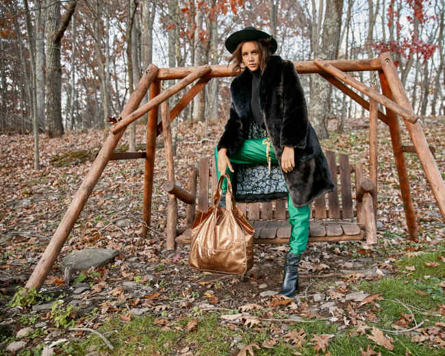 A person stands next to a wooden swing in a forested area, wearing a black fur coat and holding a large brown bag.