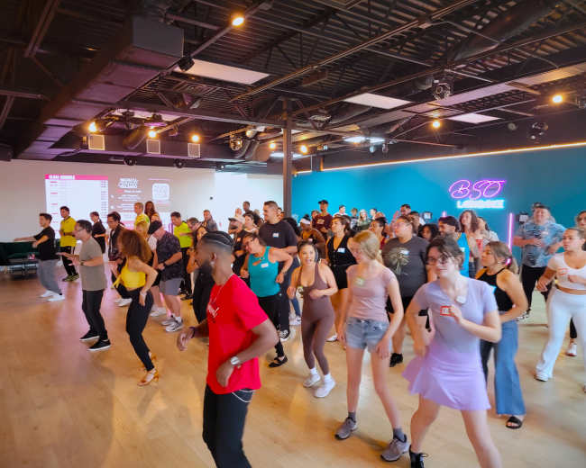 A large group of people is participating in a dance class in a studio with bright lighting and a colorful backdrop.