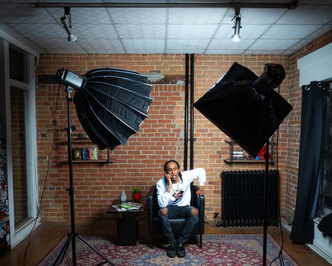 A person sits on a chair in a room with brick walls, surrounded by two softbox lights and a small table with a plant.