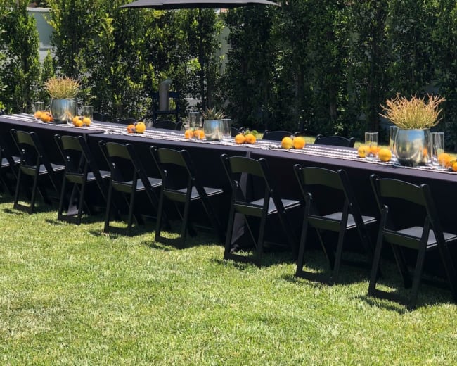 A long outdoor table set with glasses and centerpieces is positioned on a grassy lawn, surrounded by tall hedges.