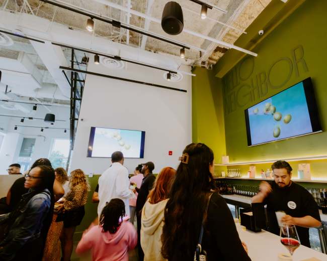 A crowd of customers gathers in a modern café with green walls and two large screens displaying visual content.