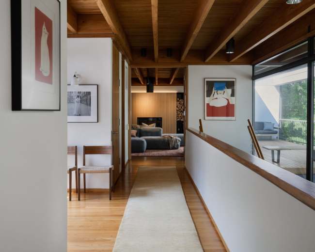 A wooden-paneled hallway features framed artwork on the walls, leading to an open living space with large windows.
