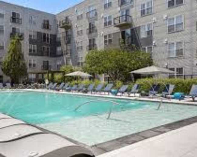 The image shows a rectangular swimming pool surrounded by lounge chairs and an apartment building with multiple floors in the background.
