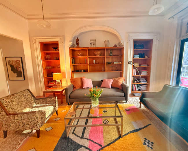 The living room features a gray sofa with pink cushions, two patterned chairs, a coffee table, a bookshelf filled with various objects, and large windows allowing natural light to enter.