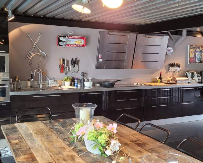 The image shows a modern kitchen with a sleek black cabinetry, a long wooden dining table with flowers, and various kitchen tools displayed on the wall.