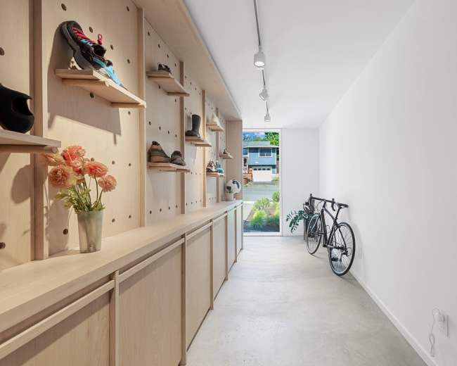 The image shows a hallway with wooden shelves displaying shoes, a countertop with flowers, and a bicycle parked against the wall near an open door.
