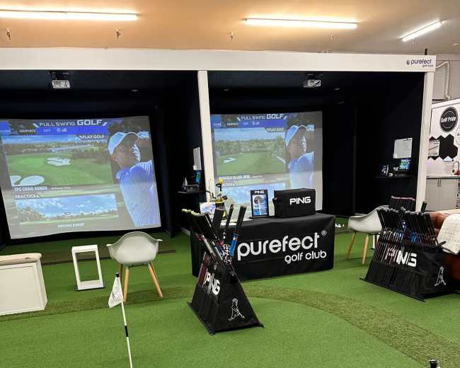 The image shows an indoor golf simulation area with putting greens, two large projection screens displaying golf courses, and various golf clubs arranged for testing.