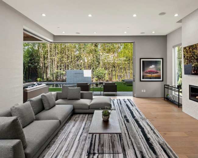 A modern living room features a large sectional sofa, a wooden coffee table, and a fireplace, with large windows showcasing a view of a bamboo garden outside.