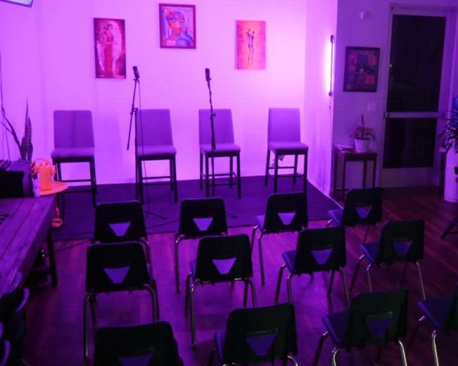 The scene shows a small indoor venue set up for an event, featuring empty chairs arranged in rows facing a small stage with two microphones and a few pieces of artwork on the walls.