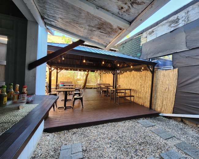 The image shows an outdoor dining area with a covered wooden gazebo, string lights overhead, and several tables arranged on a wood deck surrounded by bamboo fencing.
