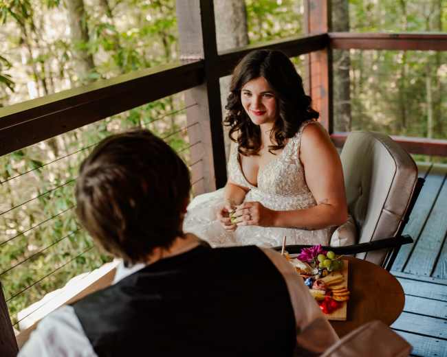 A woman in a wedding dress sits on a porch, gazing at a man seated across from her at a small table with a plate of food.